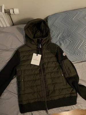 Olivgrön Moncler cardiganjacka med huva - Snygg olivgrön cardiganjacka från Moncler med vadderad front och stickade ärmar. Jackan har huva, dragkedja framtill, ribbade muddar och två fickor. Moncler-logga på ärmen och cool urban look. Perfekt för lager på lager under kyliga dagar. Stl M.