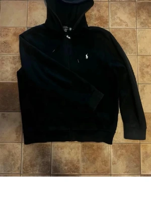 Svart hoodie med dragkedja från Polo Ralph Lauren - Säljer en svart hoodie från Polo Ralph Lauren med dragkedja framtill och vit broderad logga på bröstet. Tröjan har huva med dragsko och ribbade muddar. Perfekt för en chill och stilren look.