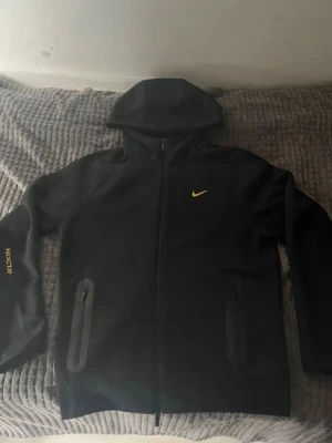 Svart Nike NOCTA hoodie med dragkedja - Snygg svart hoodie från Nike NOCTA med gul logga på bröstet och NOCTA-tryck på ärmen. 