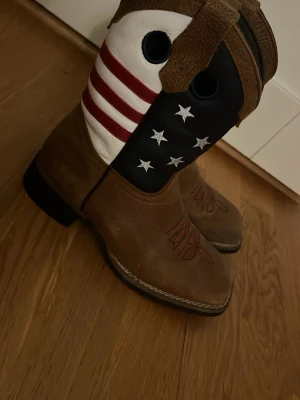 Jag säljer ett par cowboy boots💞 - Säljer ett par bruna boots som jag köpt i USA🙏🏻💞 Med fyrkantig tå och dekorativ röd söm på foten. Skaftet har amerikanska flaggans stjärnor och ränder i vitt, blått och rött🩷