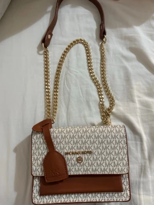 Michael Kors axelväska med kedja - Snygg axelväska från Michael Kors i beige och brunt med MK-logga över hela väskan. Väskan har guldfärgad kedjerem och detaljer i brunt läder. Praktisk ficka framtill och dekorativ tag. Perfekt för dig som vill ha en trendig och stilren väska. Den är ej äkta!