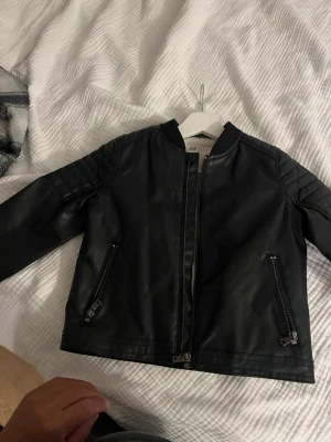 Svart skinnjacka från H&M - Snygg svart skinnjacka från H&M med raka linjer och quiltade detaljer på axlarna. Jackan har två dragkedjeförsedda fickor framtill och en stilren ståkrage utan krage. Perfekt för dig som vill ha en klassisk bikerlook.