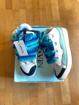 Lanvin sneakers med blå skosnören - Unika Lanvin sneakers i vitt läder med detaljer i svart och ljusblått. Skorna har extra breda, färgstarka skosnören i blå och turkosa nyanser. Klassisk låg modell med rund tå och robust sula. Perfekta för dig som vill sticka ut med din stil.