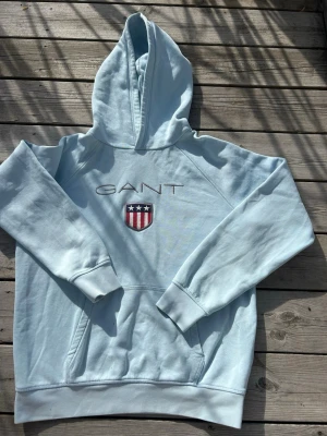 Ljusblå hoodie från GANT - Snygg ljusblå hoodie från GANT med broderad logga och amerikansk sköld på bröstet. Klassisk känguruficka framtill och ribbade muddar vid ärmslut och nederkant. Tillverkad i mjukt bomullsmaterial och har en stor huva för extra komfort.