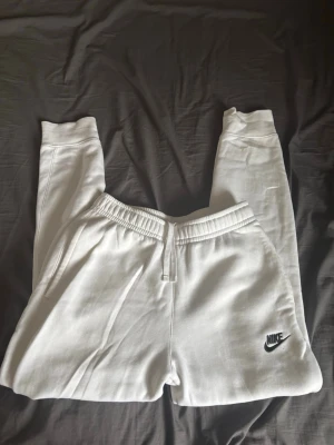 Vita mjukisbyxor från Nike - Säljer ett par vita mjukisbyxor från Nike med resår i midjan och dragsko. Byxorna har ribbade muddar vid bensluten, sidofickor och en bakficka. Nike-logga broderad i svart på vänster ben. Perfekta för chill dagar eller träning.