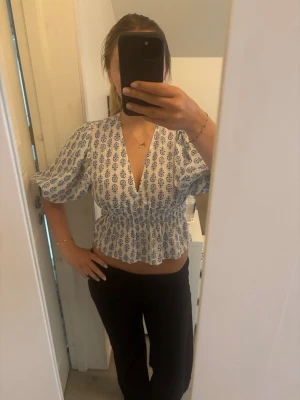 Hm blus - Viral HM blus med blått mönster, v-ringad. Blusen har volagkant nertill, korta ärmar och smockad midja som ger en feminin siluett. 