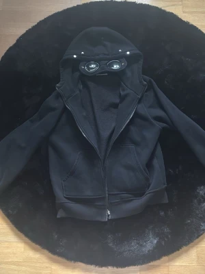 Svart hoodie med glasögon i huvan - Svart hoodie från C.P. Company med dragkedja och unika inbyggda glasögon i huvan. Hoodien har två stora fickor framtill och ribbade muddar. Perfekt för dig som vill sticka ut med en streetwear-inspirerad look.