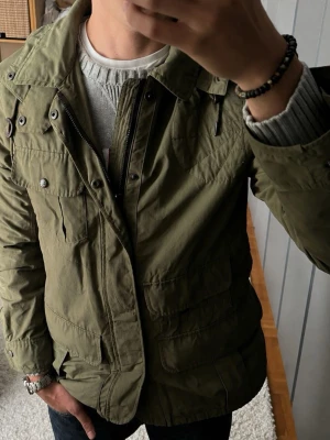 RLX Ralph Lauren fältjacka i storlek M - Mörkgrön - Riktigt snygg military/fieldjacka i tåligt bomull/nylon material med massor av fickor och fina detaljer, huva, dragkedja med tryckknappsslå över och justerbar midja.   Använd men i mycket fint skick, inga hål eller fläckar. Perfekt jacka nu till höst. Personen på bilden är 189cm, väger 78kg.  Rök och djurfritt hem. Skriv gärna om du vill ha fler bilder eller undrar något!
