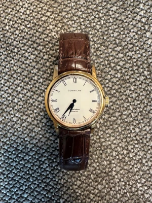 Corniche Mistral 40 limited edition 1 av 250  klocka med brunt läderband - Stilren klocka från Corniche med rund guldfärgad boett och vit urtavla med romerska siffror. Klockan har ett brunt läderband med krokodilpräglad yta och klassisk design. Perfekt accessoar för dig som gillar tidlös stil. Behöver nytt batteri. Ingen låda. 