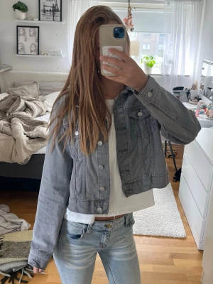 Grå jeansjacka - Helt ny snygg grå jeansjacka med klassiska bröstfickor och silverfärgade knappar. Jackan har en rak passform och långa ärmar, perfekt att slänga över en t-shirt för en avslappnad look.