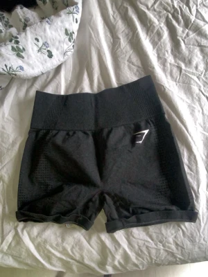 Svarta högmidjade träningsshorts - Säljer ett par svarta högmidjade träningsshorts med tight passform. Shortsen har en bred ribbad midja och diskret mönstrade sidor. Perfekta för gymmet eller löpning. Liten logga framtill på vänster ben. 