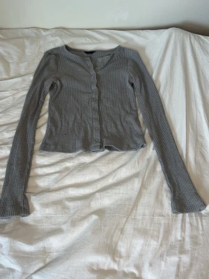 Grå ribbad cardigan med knappar - Säljer en grå ribbad cardigan med långa ärmar och knäppning framtill. Tröjan har en figurnära passform och lätt vågig kant nedtill. Perfekt att styla med jeans eller kjol för en enkel och snygg look.