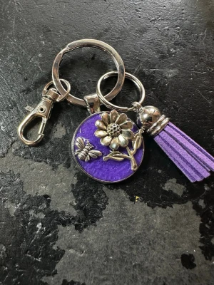 Lila nyckelring med blomma och tofs - Nyckelring med rund lila bricka dekorerad med en silverfärgad blomma och en liten fjäril. Fäst vid ringen sitter även en matchande lila tofs i mockaimitation och en metallhake för enkel fastsättning. Perfekt accessoar för att piffa upp väskan eller nyckelknippan.