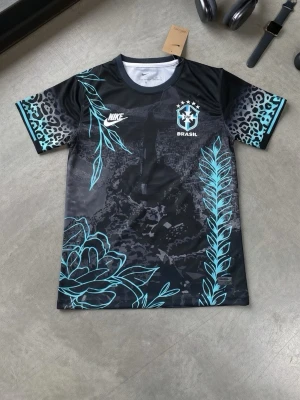 Nike Brasil svart mönstrad t-shirt - Snygg svart t-shirt från Nike med Brasil-tryck och coola turkosa blommönster framtill. Ärmarna har leopardmönster i grått och turkost. Perfekt för dig som gillar sportig och unik stil. Rund halsringning och korta ärmar.