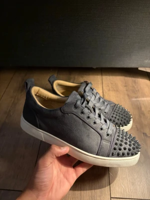 Christian loubutin skor  - Unika svarta sneakers med mattsvarta nitar på tån och ikonisk röd sula. Skorna har svarta skosnören, vit yttersula och är tillverkade i mockaliknande material. Perfekt för dig som vill sticka ut med en edgy och trendig look.