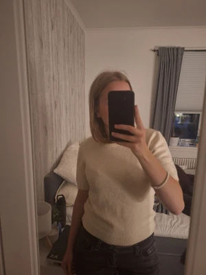 Ljusbeige stickad topp med kort ärm - Mysig ljusbeige stickad topp med korta ärmar och rund hals. Tröjan har en enkel och stilren design med ribbade kanter vid hals, ärmslut och nederkant. Perfekt att matcha till jeans eller kjol för en avslappnad look.