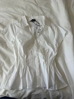 Vit blus - Säljer en blus med smickrande rynk i midjan och knappar framtill. Blusen har klassisk krage och korta ärmar. Aldrig använd och har lapparna kvar, nypris 299