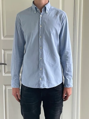 Eton skjorta - Klassisk ljusblå skjorta med button-down krage och vita knappar. Skjortan har lång ärm och rundad nederkant, perfekt för en clean och stilren look.
