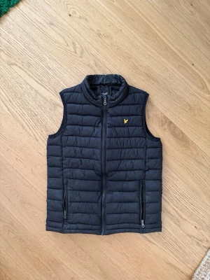 Lyle & Scott väst - Snygg svart dunväst med quiltad design och hög krage. Västen har två dragkedjeförsedda fickor framtill och en liten gul logga på bröstet. Storlek Svara och bra skick
