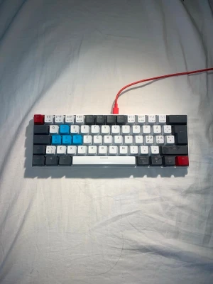 Mekaniskt tangentbord med färgglada tangenter - Säljer ett kompakt mekaniskt tangentbord med keycaps i vitt, grått, blått och rött. Tangenterna har både latinska och koreanska tecken. Tangentbordet har en röd USB-kabel och vit ram. Perfekt för gaming eller skrivande och ger en unik look till din setup.
