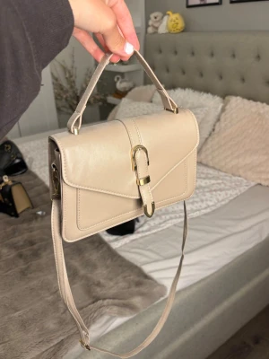 Beige axelremsväska med spänne - Supersöt beige handväska med avtagbara band så man kan välja att antingen ha den längre eller endast ha de små handtagen⭐️(Har en liten skråma där bak, se bild 2)💕
