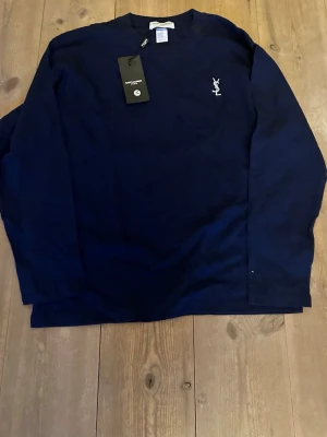 Mörkblå långärmad Saint Laurent tröja - Snygg mörkblå långärmad tröja från Saint Laurent Paris med diskret vitt logobrodyr på bröstet. Klassisk rund halsringning och mjukt material som känns skönt mot huden. Perfekt för dig som gillar stilrena plagg med exklusiv känsla. Helt oanvänd utan defekter. Fraktar inom 1-2 dagar! 