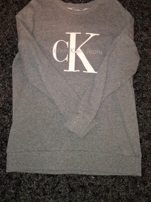 Grå sweatshirt från Calvin Klein - Mysig grå sweatshirt från Calvin Klein Jeans med stort vitt CK-tryck på bröstet. Tröjan har rund halsringning och långa ärmar. Perfekt för en avslappnad och stilren look.
