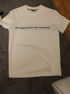 Vit t-shirt från Emporio Armani - Vit t-shirt från Emporio Armani med svart logotyptryck över bröstet. Klassisk rund halsringning och rak passform. Perfekt för en clean och stilren look.