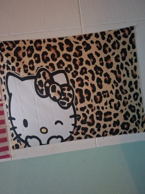 Hello Kitty poster med leopardmönster - Poster med Hello Kitty-figur i vitt med svart kontur och en leopardmönstrad rosett. Bakgrunden är beige med brunt och svart leopardmönster. Perfekt för att ge rummet en personlig och lekfull touch.