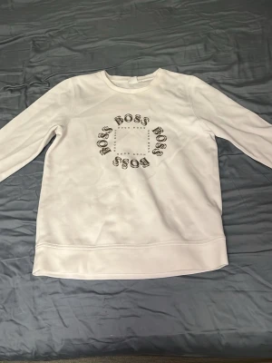 Vit sweatshirt med BOSS-tryck - Säljer en vit sweatshirt med rund hals och lång ärm. Framsidan har ett cirkelformat tryck med texten 'BOSS' flera gånger. Tröjan är i mjukt material och har en clean look utan huva. Perfekt för en chill och stilren outfit. Priset kan diskuteras vid snabb affär.