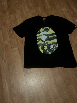 Svart BAPE t-shirt med camo-print - Säljer en svart t-shirt från BAPE med det ikoniska apansiktet i camouflage-tryck på bröstet. T-shirten har korta ärmar och en normal passform. Perfekt för dig som gillar streetwear och vill sticka ut med en unik design.