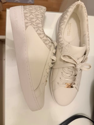 Beige sneakers från Michael Kors - Snygga beige sneakers från Michael Kors med klassiskt MK-mönster på hälen och sidorna. Skorna har vita snören, guldfärgad MK-detalj vid snörningen och är tillverkade i en mix av läder och textil. Perfekta för en trendig och stilren look.
