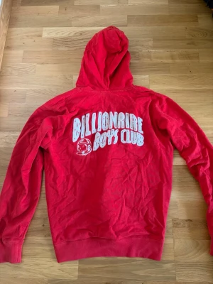 Röd Billionaire Boys Club hoodie fullzip - Snygg röd hoodie från Billionaire Boys Club med vit logga och astronaut-tryck både fram och bak. Dragkedja framtill, två fickor och klassisk huva. Perfekt för dig som vill sticka ut med streetwear-vibe. Materialet känns mjukt och bekvämt. Den är inte fadead där nere det är bara ljuset. Fullzip