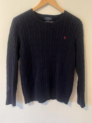 Mörkblå kabelstickad tröja Polo Ralph Lauren - Kabelstickad mörkblå tröja från Polo Ralph Lauren med rund halsringning och den klassiska röda logotypen broderad på bröstet. Tröjan har långa ärmar och ribbade muddar vid ärmslut och nederkant. Perfekt för en stilren och tidlös look.MVH Deluxe fashion 