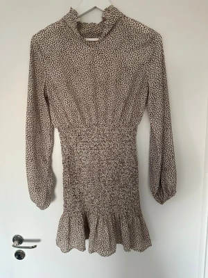 Beige prickig kortklänning - Kort klänning i beige med svart prickigt mönster. Klänningen har långärmade puffärmar, hög krage och smock i midjan samt volang nedtill. Perfekt för dig som gillar en feminin och trendig stil.