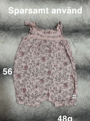 Ljusrosa blommig jumpsuit med volang - Jumpsuit i ljusrosa med blommigt mönster och volangdetaljer på axelbanden. Kortbent modell med resår vid benen och spets upptill. perfekt för en söt och romantisk look. Liten vit etikett med texten 'white' på benet.🌸Sparsamt använd. Storlek 56