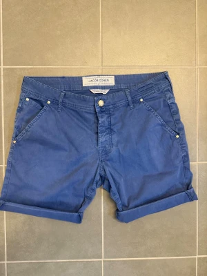 Blå shorts från Jacob Cohen, stl 34 - Snygga blå shorts från Jacob Cohen med klassisk femficksdesign och uppvikta benslut. Shortsen har normal passform, bälteshällor och knappgylf. Perfekta för varma dagar och enkel att matcha med olika stilar.