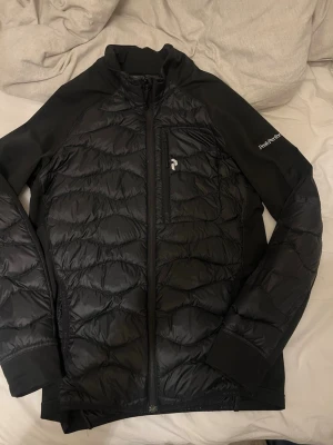 Svart pufferjacka Peak Performance - Snygg svart pufferjacka från Peak Performance med quiltat mönster och dragkedja framtill. Jackan har en bröstficka med dragkedja och logga på ärmen. Materialet är en mix av glansig och matt yta, perfekt för kalla vinterdagar.