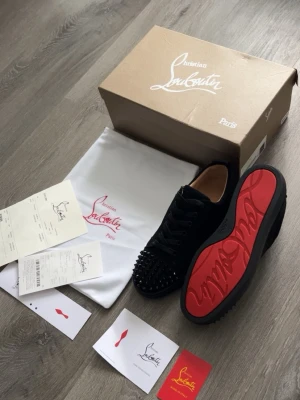 Svarta Christian Louboutin med spikar  - Svarta sneakers i mocka från Christian Louboutin med mattsvarta nitar på tån och den ikoniska röda sulan. Skorna har snörning, låg profil och rund tå för en lyxig och edgy look.