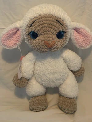 Handvirkat lamm med blå ögon - Supergulligt mjukisdjur i form av ett lamm med stora blå ögon, vit fluffig kropp och beige ansikte, tassar och öron. Insidan av öronen är rosa och nosen är broderad. Tillverkad i mjuk plysch, perfekt att krama eller ha som dekoration. 35 cm hög