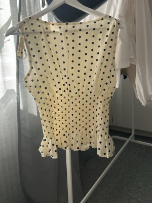 Prickig ärmlös topp med volang - Söt ärmlös topp i ljusgul med svarta prickar. Toppen har smock i midjan för en figurnära passform och volangkant nedtill. Axelbanden knyts på axlarna för en extra detalj. Perfekt för en trendig och somrig look. ljuset gör att den ser gul ut 