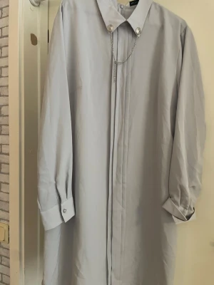 Ljusblå långärmad tunika/blus med kedjedetalj - Snygg ljusblå blus i längre modell med klassisk krage och silverfärgad kedjedetalj mellan kragspetsarna. Dolda knappar framtill och långa ärmar med manschett. Perfekt för en clean och stilren look.