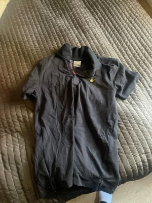 Svart piké från Lyle & Scott - Säljer en svart pikétröja från Lyle & Scott med klassisk krage och knappslå med röd och blå detalj. Tröjan har korta ärmar och den ikoniska gula logon broderad på bröstet. Perfekt till en avslappnad men ändå stilren look.