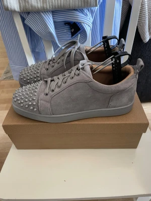 Gråa louboutin - Säljer ett par grå sneakers i mocka-look med coola nitar över hela tån. Skorna har snörning och en låg profil som ger en clean och modern vibe. Perfekta för dig som vill sticka ut med din stil och gillar detaljer som syns. Skorna är änvänd 1 gång bara och de kommer med shoe box.