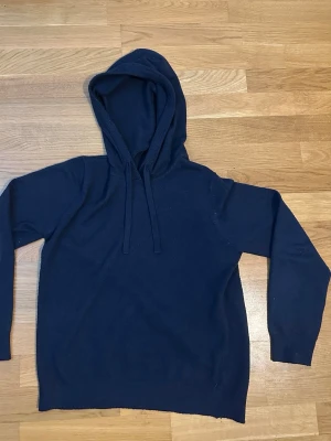 Marinoull tröja - Mörkblå hoodie från RYVLS Stockholm med klassisk huva och dragsko. Tillverkad i mjukt stickat material som känns skönt mot huden. Perfekt för dig som gillar en enkel och stilren look. Långa ärmar och ribbade muddar för extra komfort.