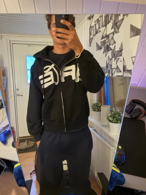 Svart hoodie med dragkedja från Arte - Säljer en svart hoodie från Arte med stor vit logga över bröstet. Hoodien har dragkedja framtill, två fickor och en klassisk huva med snören. Perfekt för dig som gillar streetwear och vill ha en stilren men ändå iögonfallande look.