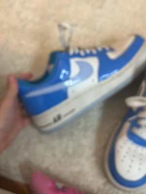 Blåvita Nike Air Force 1 sneakers - Säljer ett par blåvita Nike Air Force 1 sneakers med klassisk låg siluett. Skorna har vit sula med 'AIR'-detalj, blått och vitt läder samt vita skosnören. Perfekta för dig som vill ha en ikonisk och stilren sneaker till din outfit.