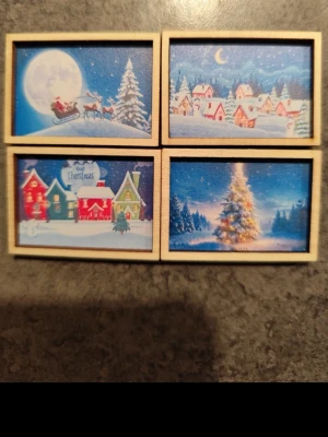 Små juliga minitavlor i trä - Fyra små rektangulära minitavlor med julmotiv, perfekta som dekoration. Motiven visar snötäckta byar, tomte med släde, julgran och färgglada hus. Tavlorna har träram och färgstarka vinterbilder i blått, rött och vitt.