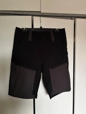 Svarta och grå funktionsshorts - Snygga funktionsshorts i svart och mörkgrått med praktiska dragkedjefickor på sidorna. Shortsen har bälteshällor och elastisk midja baktill för extra komfort. Perfekta för utomhusaktiviteter och äventyr.