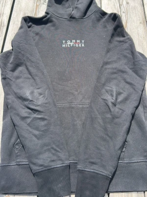 Svart hoodie från Tommy Hilfiger - Snygg svart hoodie från Tommy Hilfiger med logga på bröstet. Tillverkad i en mjuk ekologisk bomullsblandning. Klassisk huva och känguruficka framtill. Perfekt för en chill och stilren look.
